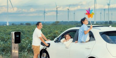 İlerici mutlu aile kavramı elektrikli araçla rüzgar çiftliğinde eğleniyor. Rüzgar türbini jeneratöründen gelen temiz yenilenebilir enerji ile şarj edilen elektrikli araç..