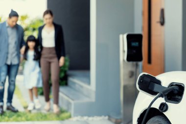 Focus closeup electric vehicle recharging battery from home electric charging station with blurred family in background. Renewable clean energy car for progressive eco awareness lifestyle concept.