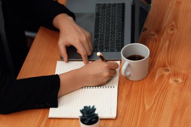 Closeup hand of secretary writing on notebook with laptop and coffee at her desk in harmony office. Businesswoman sitting on her workstation taking note in the meeting for financial strategy planning.