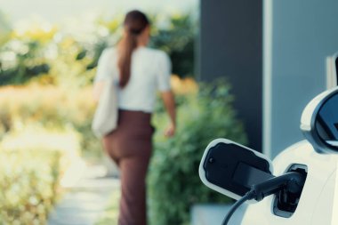 Focus EV charger plugged into EV car at home charging station with blurred background of progressive woman walking in background. Elective vehicle powered by clean energy for eco-friendly concept.