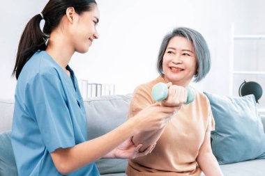 Caregiver helping contented senior woman exercise with dumbbell at home. Rehab for elder, senior medical care.