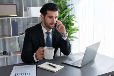 Modern ofis çalışma alanında çalışan bir işadamı elinde bir fincan kahveyle telefon görüşmelerini cevaplarken ya da çalışanları yönetirken. Coşkulu