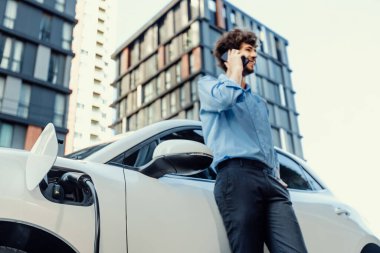 Progressive businessman talking on the phone, leaning on electric car recharging with public EV charging station, apartment condo residential building on the background as green city lifestyle.