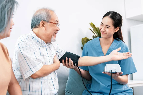 stock image An elderly man having a blood pressure check by his personal caregiver with his wife sitting next to him in their home.