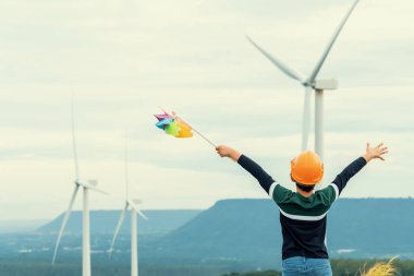 Progressive young asian boy playing with wind pinwheel toy in the wind turbine farm, green field over the hill. Green energy from renewable electric wind generator. Windmill in the countryside concept