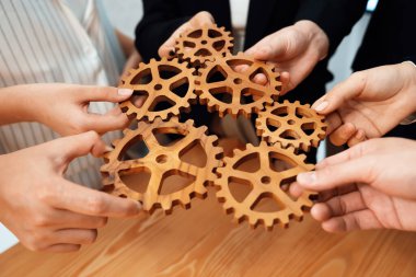 Closeup hand holding wooden gear by businesspeople wearing suit for harmony synergy in office workplace concept. Group of people hand making chain of gears into collective form for unity symbol.