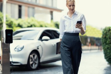 İş kadını tablet kullanıyor, halka açık otoparkta elektrikli aracını şarj ederken yürüyor. Teknolojinin ilerici yaşam tarzı ve EV otomobilinin ekolojik kaygısı