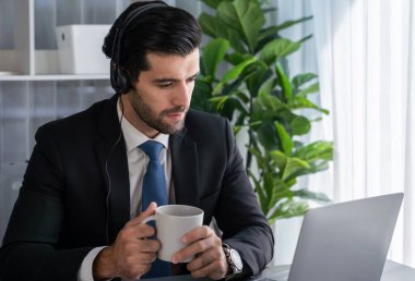 Çağrı merkezi operatörünün müdürü bilgisayarla uğraşırken masasında kahvesiyle oturuyor. Kulaklık takan ve siyah takım giyen bir ofis çalışanı müşteri desteği ya da tele pazarlama üzerinde çalışıyor. coşkulu