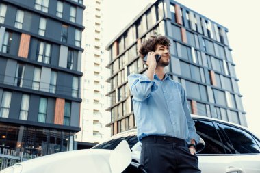 Progressive businessman talking on the phone, leaning on electric car recharging with public EV charging station, apartment condo residential building on the background as green city lifestyle.
