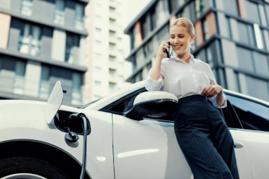 Closeup progressive businesswoman talking on smartphone, leaning electric car at charging station with residential condo apartment in background. Eco friendly rechargeable EV car concept.