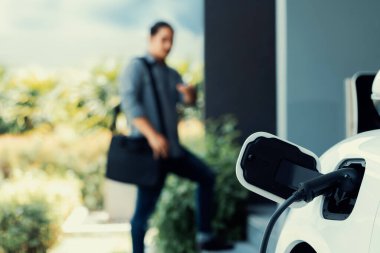 Focus electric car charging at home charging station with blurred progressive man walking in the background. Electric car using renewable clean for eco-friendly concept.