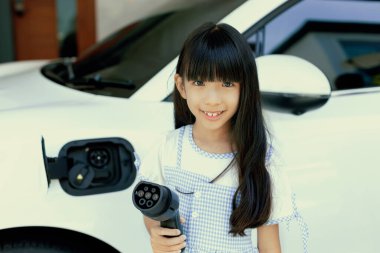 A playful girl holding and pointing an EV plug, a home charging station providing a sustainable power source for electric vehicles. Concept of progressive new generation with ecological awareness.