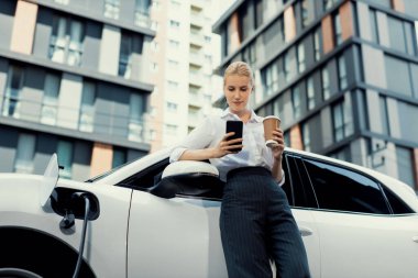 Focus businessman using phone, leaning on electric vehicle, holding coffee with blurred city residential condo buildings in background as progressive lifestyle by renewable and sustainable EV car.