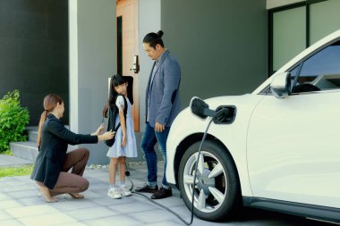 Progressive young parents and daughter with electric vehicle and home charging station. Green and clean energy from electric vehicles for healthy environment. Eco power from renewable source at home.