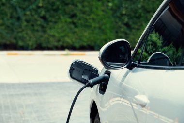 Closeup electric vehicle plugged in with EV charger at public charging station powered by renewable clean energy with residential building in background as progressive concept.