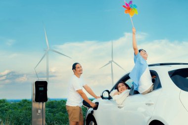 Concept of progressive happy family holding windmill toy and relax at wind farm with electric vehicle. Electric vehicle driven by clean renewable energy from wind turbine generator to charger station.