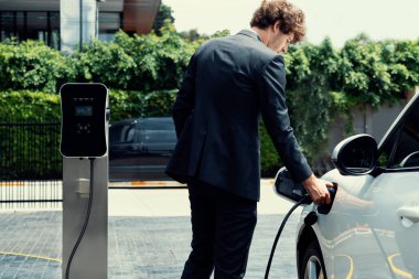 Modern şehir yerleşim bölgesindeki kamu şarj istasyonunda elektrikli araba şarj eden yenilikçi bir işadamı. Çevre dostu şarj edilebilir araba alternatif temiz enerjiyle çalışıyor..