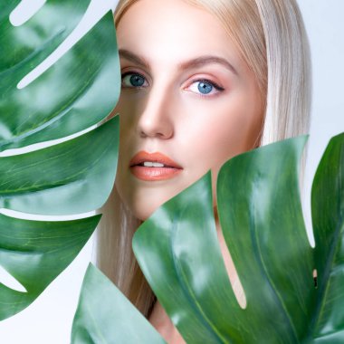 Closeup facial portrait personable woman with perfect smooth makeup holding green monstera leaves and cover her face as natural healthy skincare treatment. Tropical nature and beauty concept