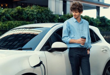 Progressive eco-friendly concept of parking EV car at public electric-powered charging station in city with blur background of businessman leaning on recharging-electric vehicle with coffee.