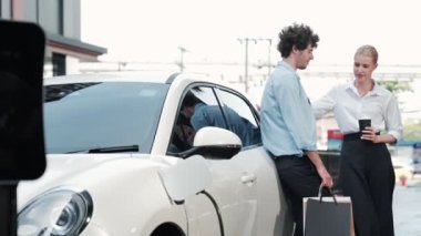 İlerici iş adamı ve iş kadını şehir merkezinde dolaşmadan önce şarj istasyonuna bağlı elektrikli arabaya yaslanıyor. Çevre dostu şarj edilebilir araba temiz enerjiyle çalışıyor..