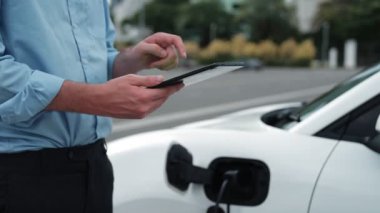 Takım elbiseli, hırslı bir işadamı elektrik kablosu ve yenilenebilir enerji enerjili bir elektrikli araçla şarj istasyonunda dururken elektrikli aracına yaslanıyor..