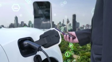 İlerleyen yeşil şehir ESG sembolü, elektrikli araçla birlikte. İş adamı EV arabalarını şarj etmek için şarj aletini şarj ediyor. Şehirdeki şarj istasyonunda ekolojik ve yeşil semboller var..