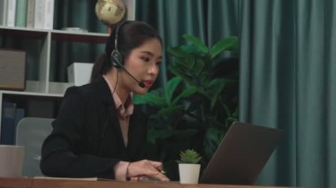 Kulaklık ve mikrofon takan iş kadını masasında çalışıyor ve dizüstü bilgisayar kullanıyor. Uzaktan kumandalı online toplantı için dizüstü bilgisayar kullanan hevesli kadın çalışanlar, ofiste iş videosu konferansı.