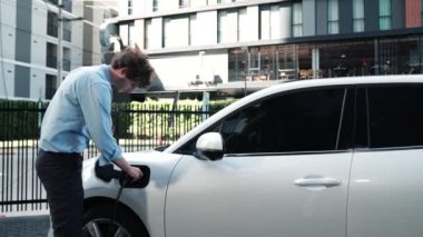 İlerici işadamı, şehir merkezinde dolaşmadan önce şarj cihazından elektrikli arabasına şarj prizi takıyor. Çevre dostu şarj edilebilir araba, sürdürülebilir ve temiz enerjiyle çalışıyor..
