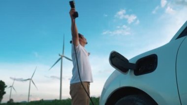 Elektrikli arabasıyla ilerici bir adam, EV arabası gelecekteki sürdürülebilir enerji kavramı olarak rüzgar türbiniyle yeşil alandaki enerji şarj etme istasyonundan enerji yüklüyor. Enerji üreteci olan elektrikli bir araç..