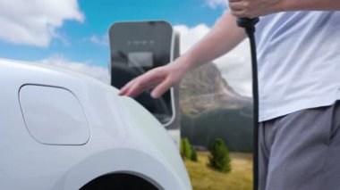 İlerici adam şarj noktasında elektrikli araba yüklüyor, daha temiz bir çevre için temiz ve yenilenebilir enerjiyle çalışan bir EV ile doğada ve dağlarda seyahat ediyor..