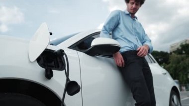 Takım elbiseli, hırslı bir işadamı elektrik kablosu ve yenilenebilir enerji enerjili bir elektrikli araçla şarj istasyonunda dururken elektrikli aracına yaslanıyor..