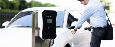 Progressive eco-friendly concept of focus parking EV car at public electric-powered charging station in city center with blur businessman walking in the background.