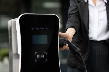 Focus EV charger plug and electric car at public charging station with blur progressive businesswoman holding charger and apartment condo building in background. Eco friendly electric vehicle concept.