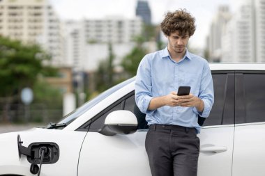 Progressive businessman talking on the phone, leaning on electric car recharging with public EV charging station, apartment condo residential building on the background as green city lifestyle.