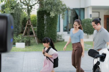 Progressive young parents and daughter with electric vehicle and home charging station. Green and clean energy from electric vehicles for healthy environment. Eco power from renewable source at home.