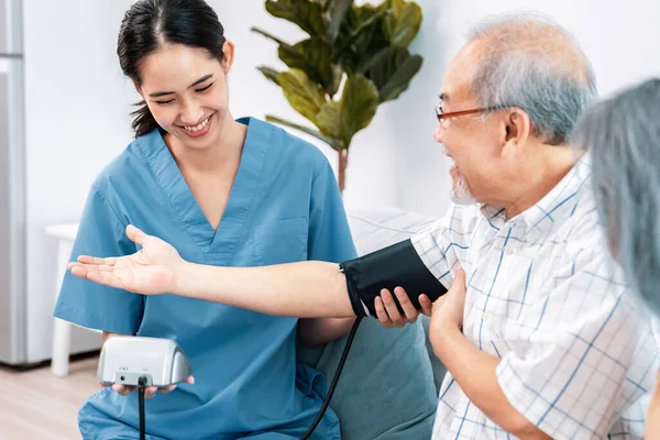 stock image An elderly man having a blood pressure check by his personal caregiver with his wife sitting next to him in their home.