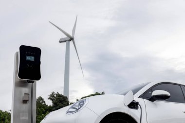 Progressive combination of wind turbine and EV car, future energy infrastructure. Electric vehicle being charged at charging station powered by renewable energy from wind turbine in the countryside.