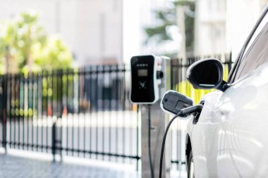 Focus closeup electric vehicle plugged in with EV charger device from blurred background of public charging station powered by renewable clean energy for progressive eco-friendly car concept.
