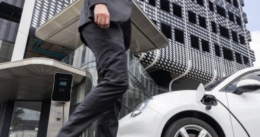 Closeup progressive businessman with electric car recharging at public charging station with background of city residential building. Eco friendly car powered by alternative clean energy.