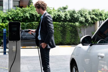 Modern şehir yerleşim bölgesindeki kamu şarj istasyonunda elektrikli araba şarj eden yenilikçi bir işadamı. Çevre dostu şarj edilebilir araba alternatif temiz enerjiyle çalışıyor..