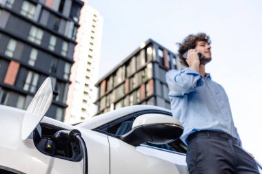 Progressive businessman talking on the phone, leaning on electric car recharging with public EV charging station, apartment condo residential building on the background as green city lifestyle.