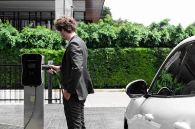 Modern şehir yerleşim bölgesindeki kamu şarj istasyonunda elektrikli araba şarj eden yenilikçi bir işadamı. Çevre dostu şarj edilebilir araba alternatif temiz enerjiyle çalışıyor..