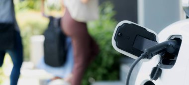 Focus closeup electric vehicle recharging battery from home electric charging station with blurred family in background. Renewable clean energy car for progressive eco awareness lifestyle concept.