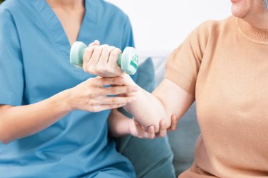 Caregiver helping contented senior woman exercise with dumbbell at home. Rehab for elder, senior medical care.