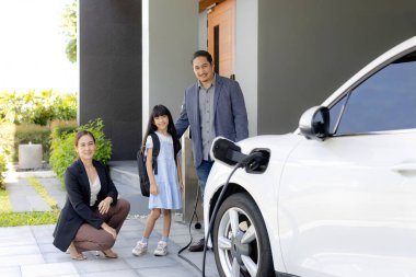 Progressive young parents and daughter with electric vehicle and home charging station. Green and clean energy from electric vehicles for healthy environment. Eco power from renewable source at home.