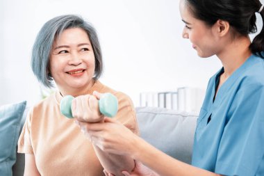 Caregiver helping contented senior woman exercise with dumbbell at home. Rehab for elder, senior medical care.