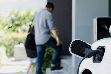 Arka planda bulanık baba ve kızının yürüdüğü şarj istasyonunda elektrikli araba şarj etmeye odaklan. Sağlıklı çevre yaşam tarzı konsepti için yenilikçi yeşil ve temiz enerji aracı.