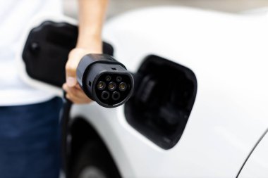 Progressive new alternative clean energy vehicle and electric charging device concept. Focus closeup hand holding and pointing EV charger plug at camear from electric charging station at camera