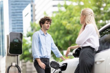 Progressive businessman and businesswoman with electric car parking and connected to public charging station before driving around city center. Eco friendly rechargeable car powered by clean energy.