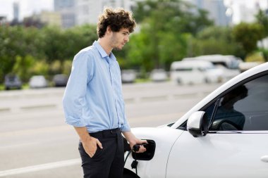 Progressive businessman insert charger plug from charging station to his electric vehicle with apartment condo building in background. Eco friendly rechargeable car powered by sustainable energy.